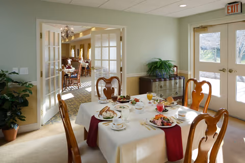 A bright dining room with a table set for four, wooden chairs, and French doors opening into a larger dining area.