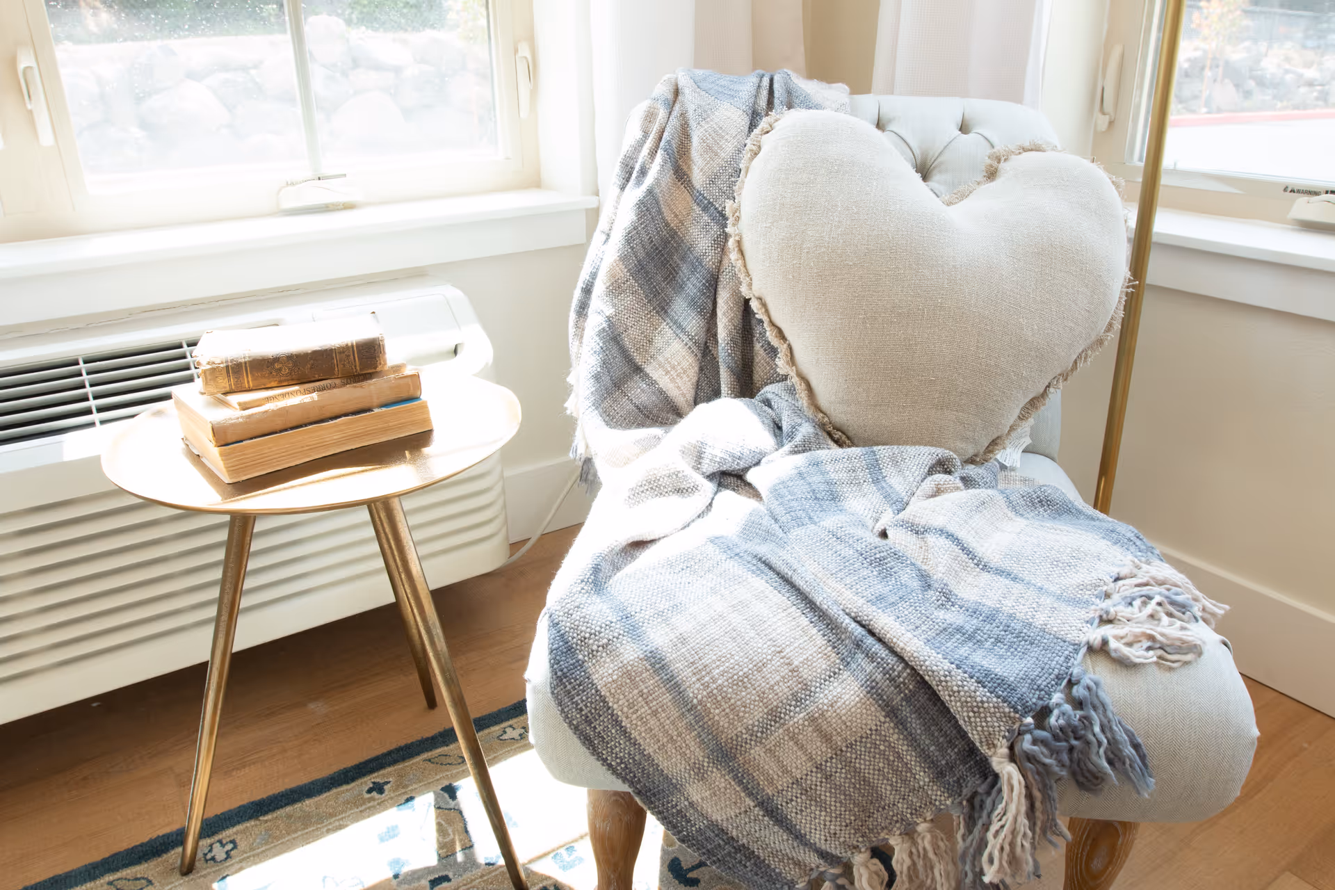 A cozy corner with a cushioned chair draped with a blue and beige plaid throw blanket and a heart-shaped pillow. Next to the chair is a small round wooden side table with a stack of old books. The scene is lit by natural light coming through two windows behind the chair.