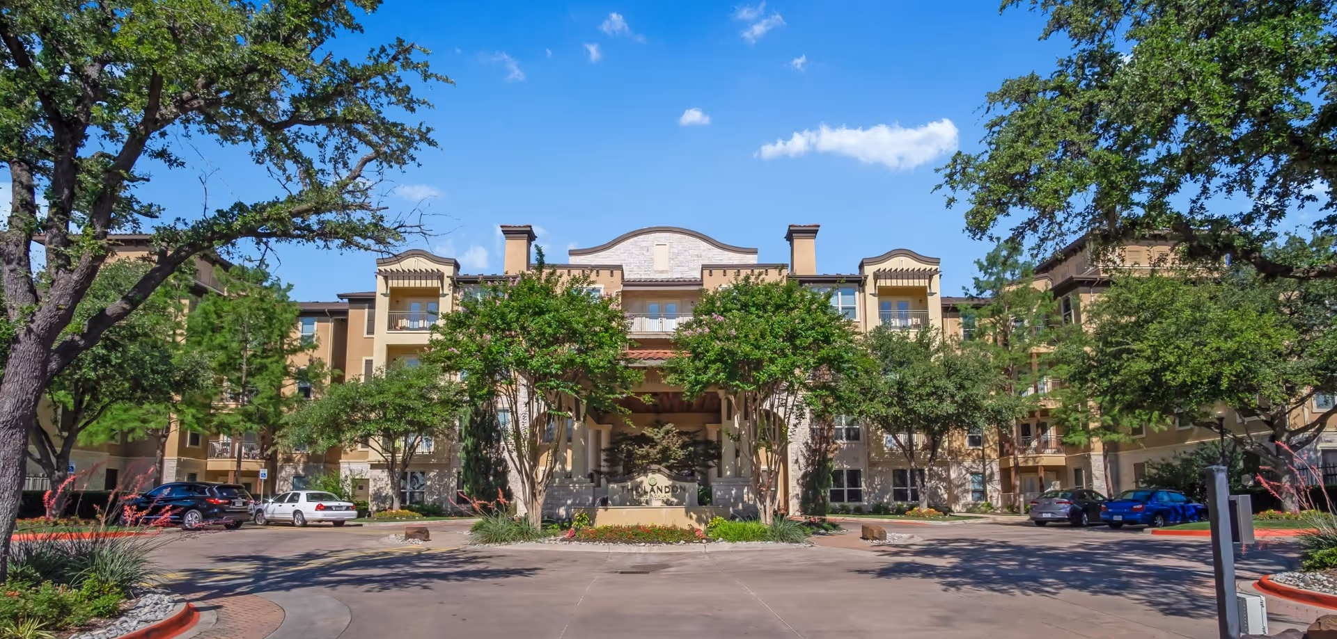 Front exterior of a multi-story senior living building with a circular driveway, trees, and parked cars under a blue sky.