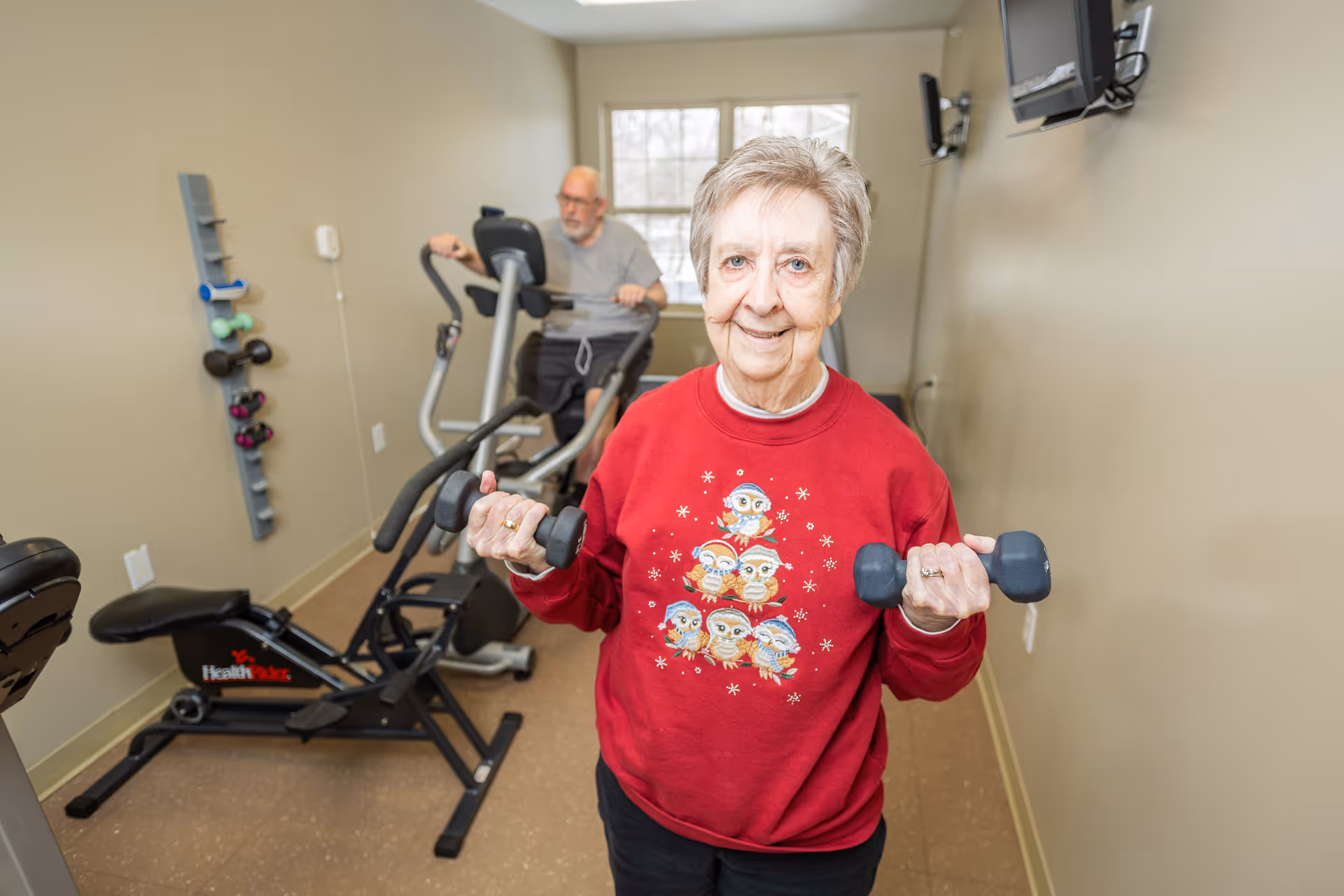 An elderly woman in a red sweater with owl designs is holding dumbbells and smiling at the camera in a small exercise room. In the background, an elderly man is using an elliptical machine. The room has beige walls, a window, and mounted televisions.