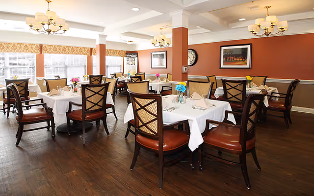 Bright dining room with several tables draped in white linens and wooden chairs under chandeliers.