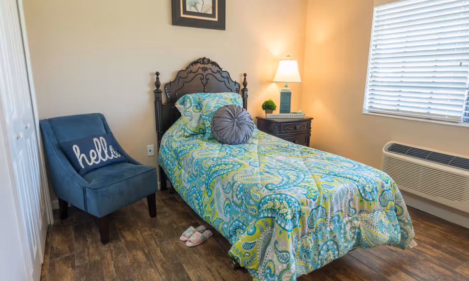 A cozy bedroom with a single bed featuring a colorful paisley-patterned bedspread in shades of green, blue, and yellow. Next to the bed is a dark wooden nightstand with a lamp, a small plant, and a book. A blue upholstered chair with a pillow that says 'hello' is positioned near a white closet door. The room has wooden flooring and a window with white blinds, and a wall-mounted air conditioning unit below the window.