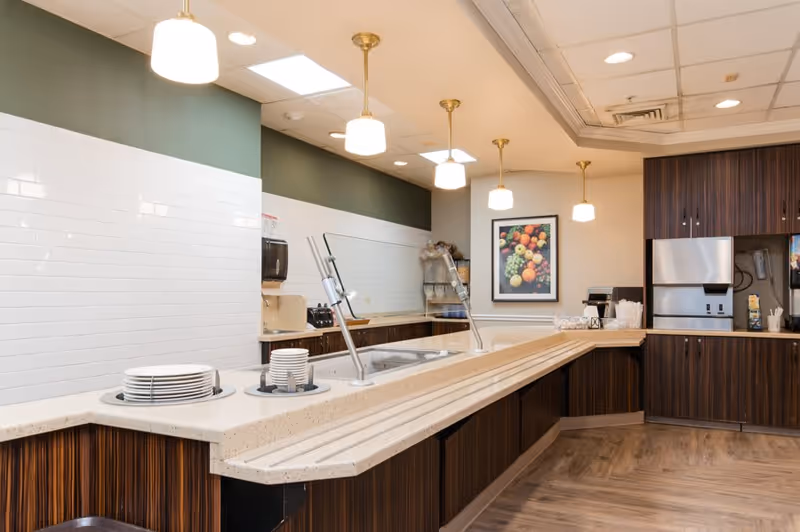 A clean and modern kitchen area with a long countertop featuring a built-in serving station with plates and bowls stacked on one side. The back wall has dark wood cabinets, a stainless steel appliance, and a framed picture of assorted fruits. The ceiling has recessed lighting and hanging pendant lights.