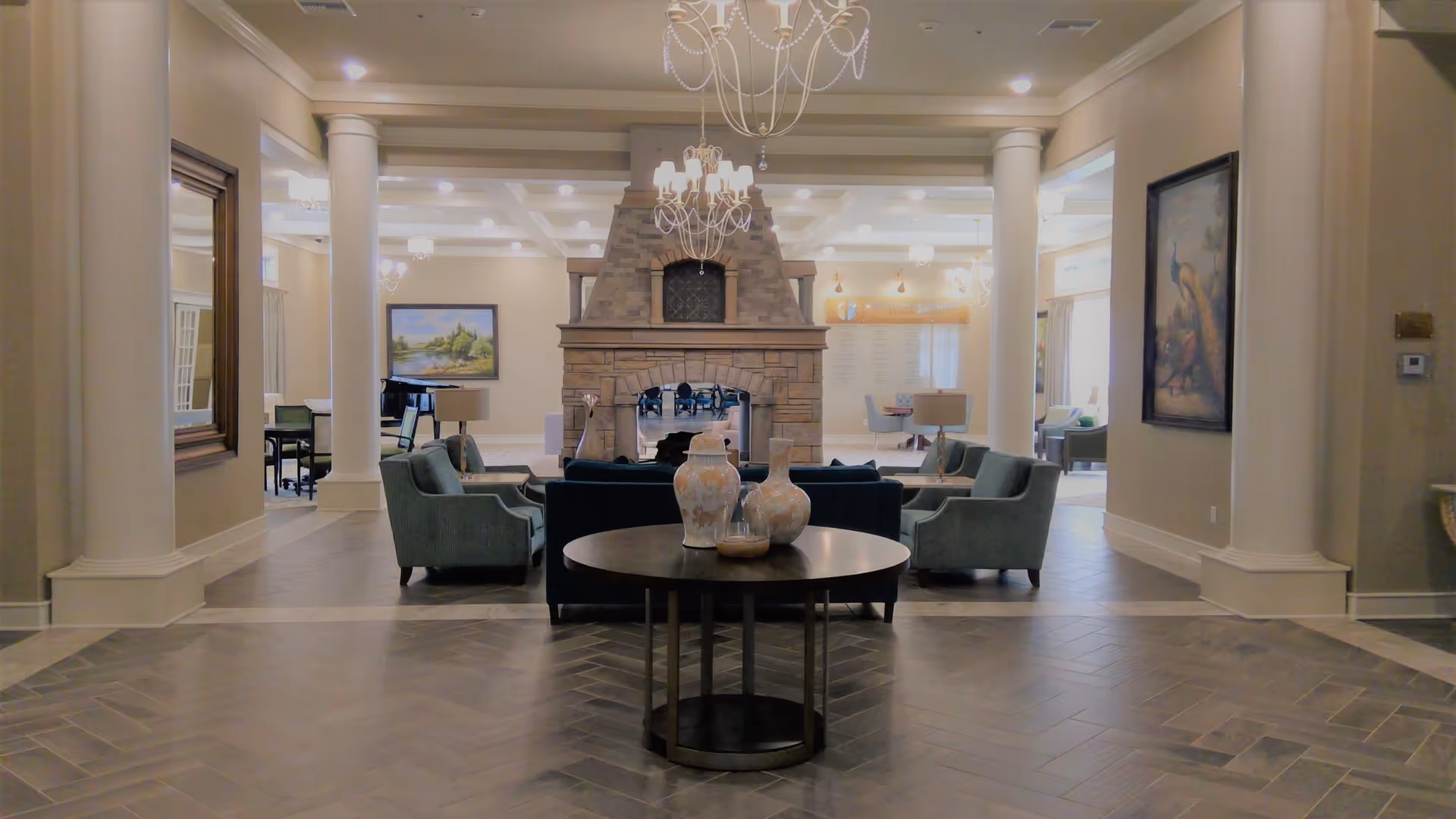 A spacious and elegant living room area with a large stone fireplace in the center. The room features multiple comfortable armchairs and sofas arranged around the fireplace. There are decorative vases on a round table in the foreground, chandeliers hanging from the ceiling, and framed artwork on the walls. The room has tall white columns and large windows allowing natural light to enter.