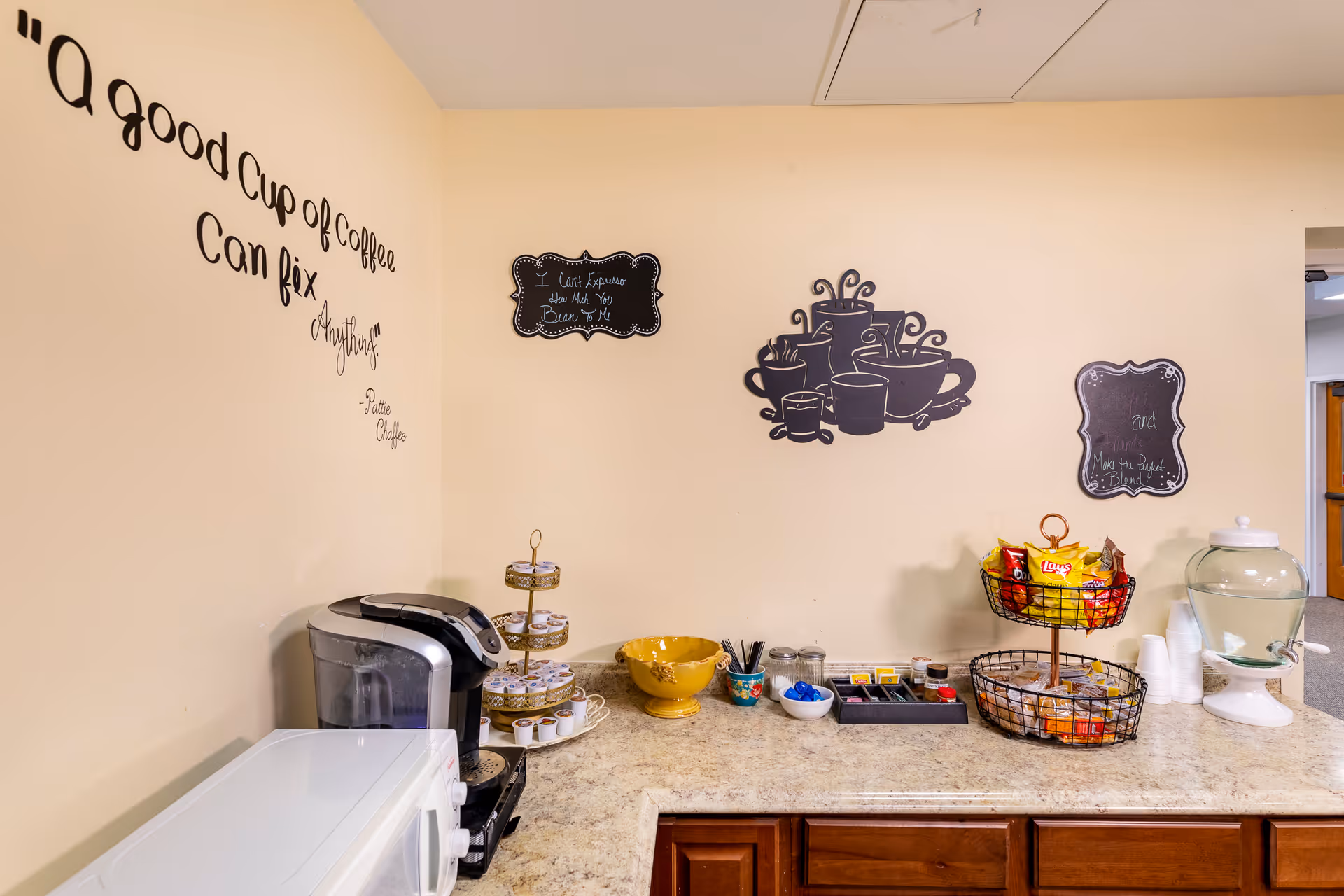 A coffee and snack station with a coffee maker, microwave, various snacks in baskets, condiments, and a water dispenser on a countertop. The wall has decorative coffee-themed signs and a quote that reads, 'A good cup of coffee can fix anything.'