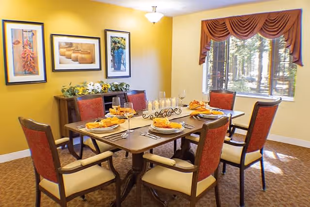 A dining room with a wooden table set for six people. The table has plates, silverware, wine glasses, and orange napkins. Six chairs with red upholstery surround the table. The walls are painted yellow and decorated with three framed pictures. A window with a brown valance shows a view of trees outside.