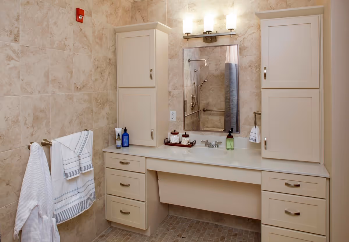 A well-lit bathroom featuring a modern vanity with a sink, storage cabinets, and a large mirror. The walls are tiled with a light stone pattern, and there are neatly arranged toiletries on the counter. Two white towels hang on a rack, adding a touch of comfort to the space.
