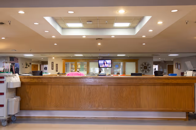 Long wooden reception desk in a senior living facility lobby with a TV screen and recessed ceiling lights above.