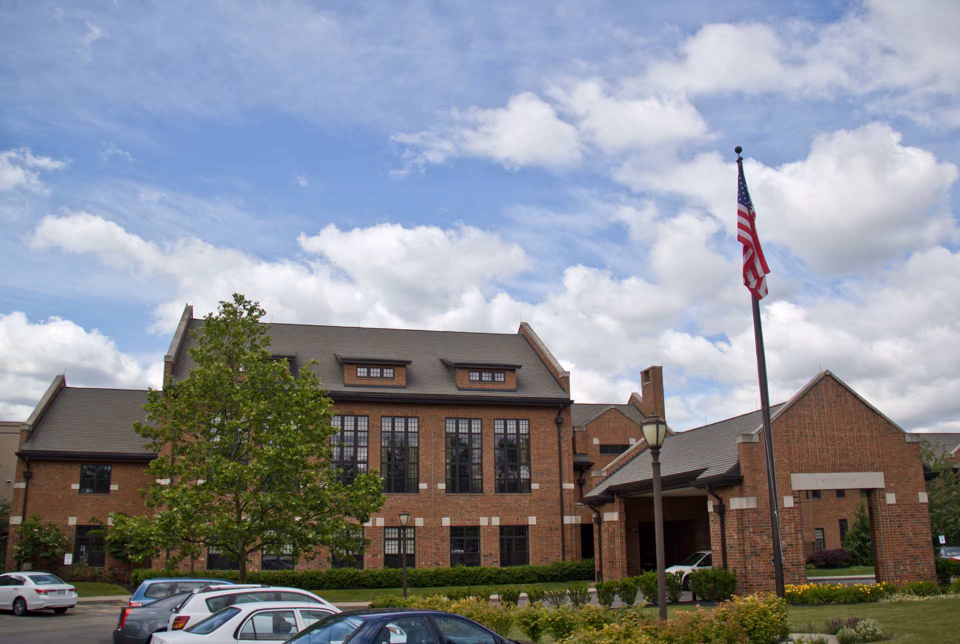 A brick senior living facility with large windows, surrounded by greenery and an American flag.