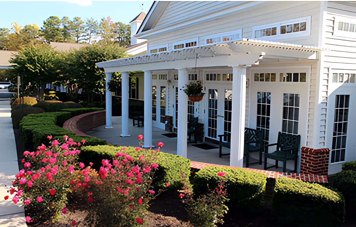 Front porch of a white senior living building with columns, chairs, and landscaped flower beds with pink blooms.