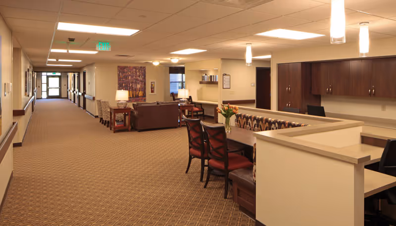 Interior view of a senior living facility hallway with a seating area featuring chairs, a sofa, tables, and lamps. The hallway is carpeted with patterned flooring and has beige walls with wooden trim. There is a reception or desk area with chairs and cabinets on the right side, and an exit sign is visible at the end of the hallway.