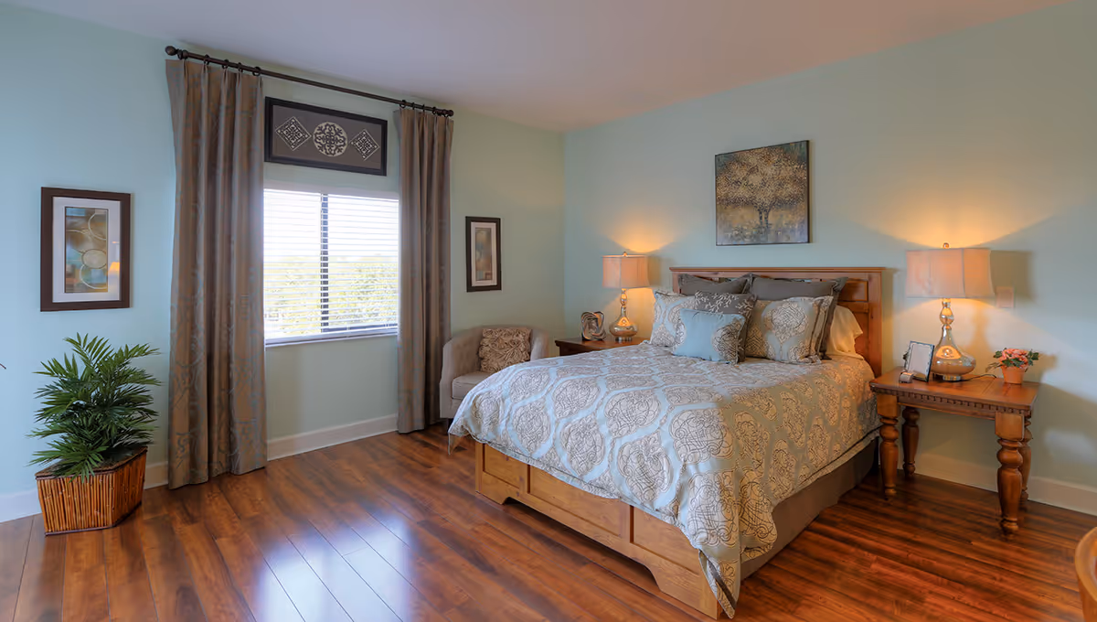 Well-lit bedroom with a wooden bed dressed in patterned bedding, two bedside tables with lamps, a window with curtains, a chair, and a potted plant on hardwood floors.