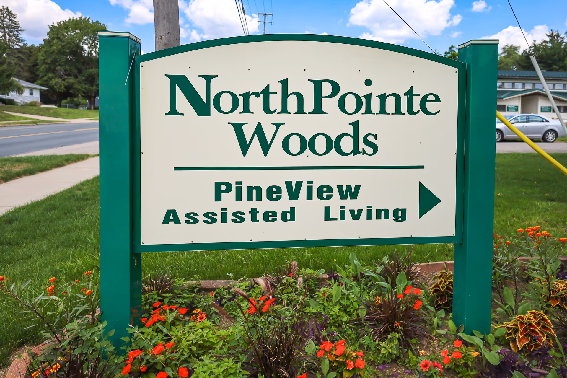 Outdoor sign for NorthPointe Woods with an arrow pointing to PineView Assisted Living, surrounded by green grass and colorful flowers, with a street and buildings in the background under a partly cloudy sky.