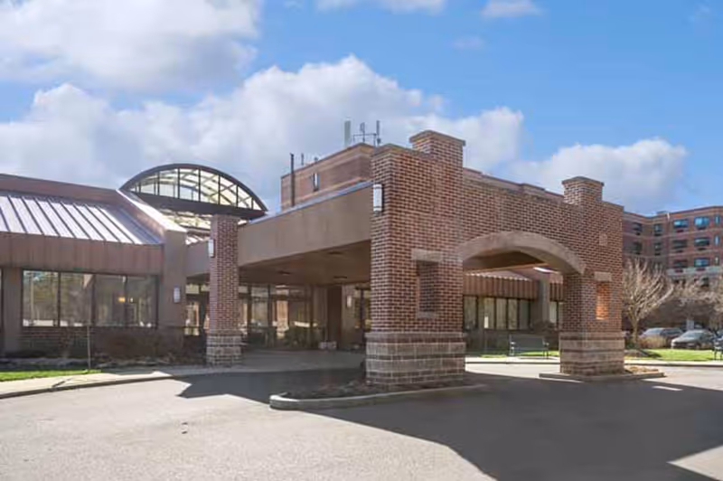 Exterior view of a senior living facility named Leisure Park showing a brick entrance canopy with an archway, large windows, and a clear blue sky with some clouds.