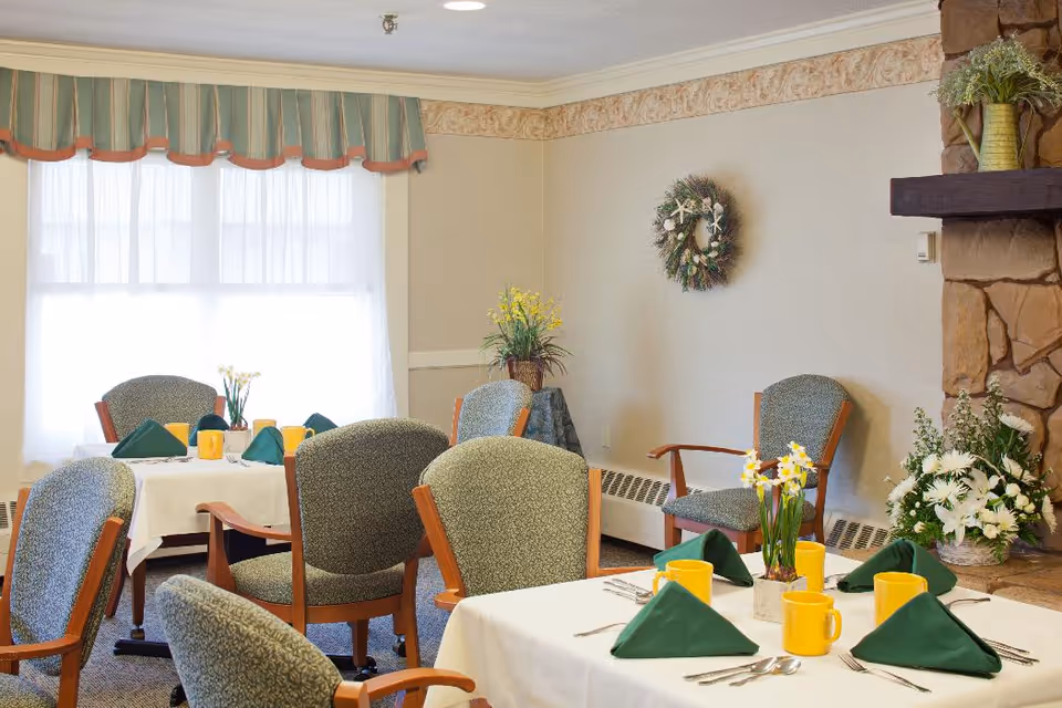 A dining room with tables set for a meal, featuring white tablecloths, green folded napkins, yellow mugs, and floral centerpieces. The room has upholstered chairs, a window with sheer curtains and a valance, a stone fireplace with floral arrangements, and a decorative wreath on the wall.