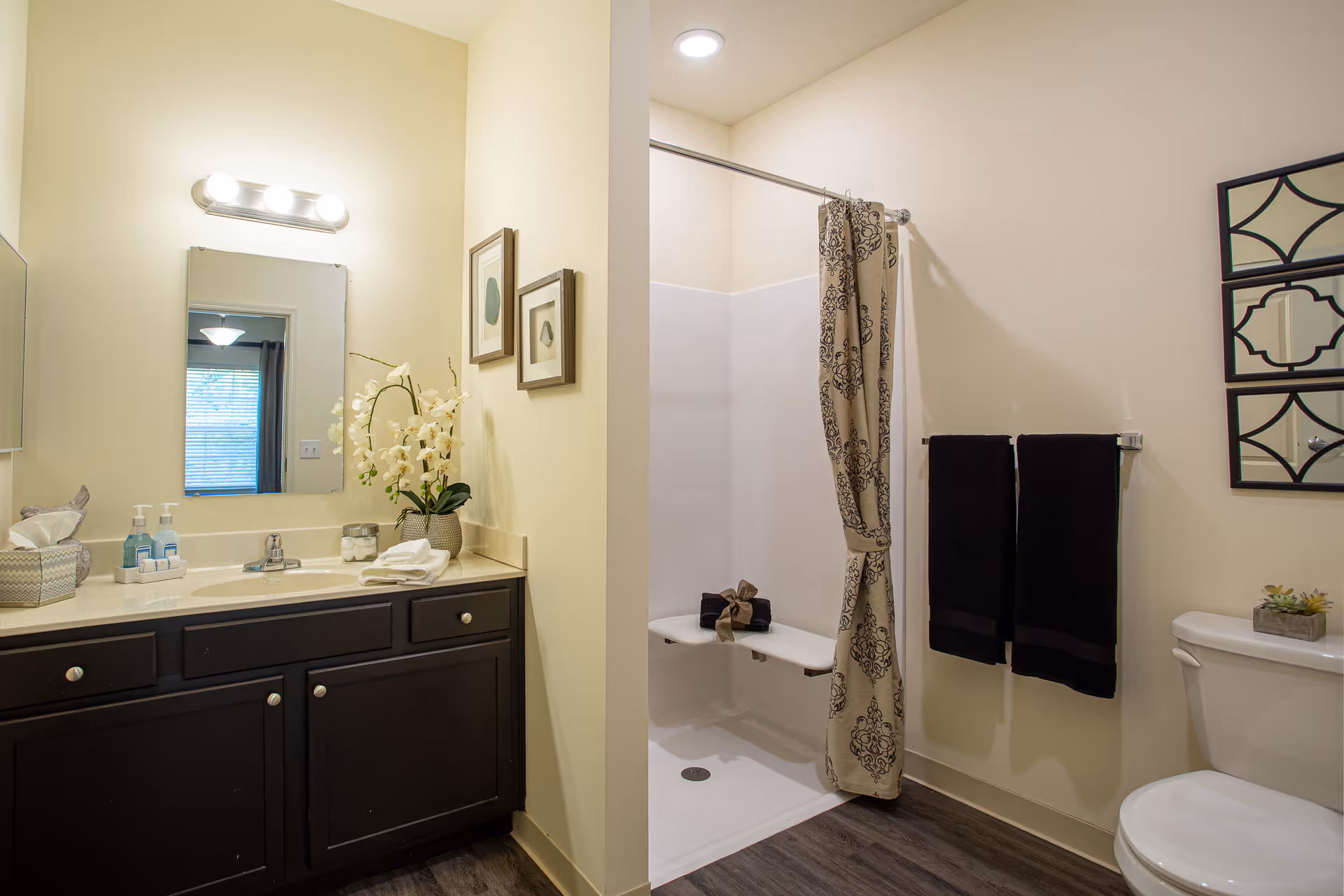 A clean and well-lit bathroom featuring a dark wood vanity with a sink, a mirror above it, and decorative items including a tissue box, soap dispensers, and a vase with white flowers. To the right, there is a walk-in shower with a patterned shower curtain tied back, a built-in shower seat with folded towels, two black towels hanging on a rack, and a toilet with a small plant on the tank.