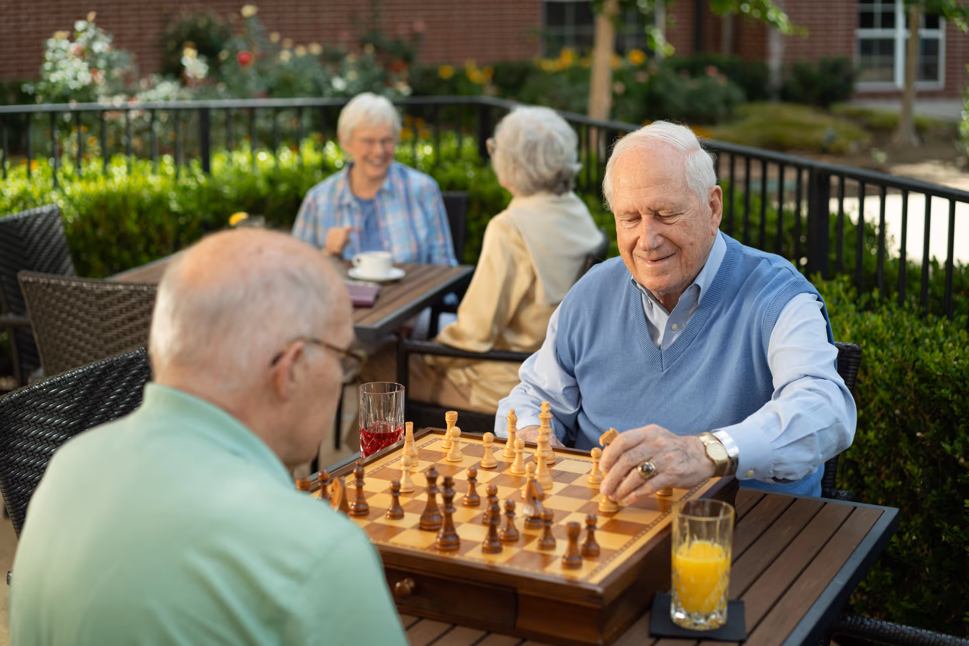 Two elderly men playing chess at a wooden table outdoors with drinks beside them, while two elderly women sit at another table in the background, enjoying conversation in a garden patio area with greenery and a brick building visible.