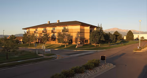 Three-story senior living building with balconies, trees, and landscaped lawn beside a road and mountains in the background.