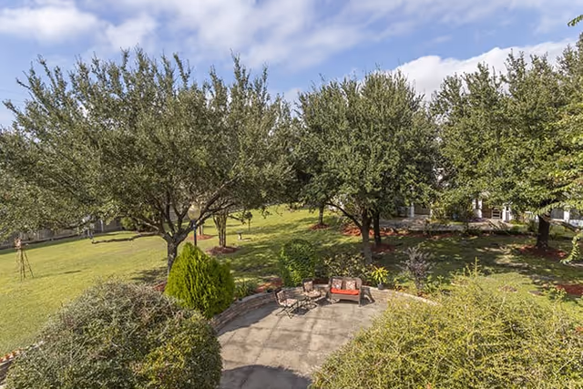 Outdoor patio area with chairs and a small table surrounded by green bushes and trees under a partly cloudy sky in a garden setting.