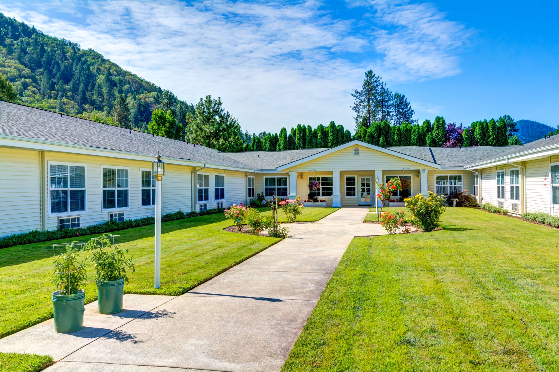 Exterior view of Morrow Heights Assisted Living facility showing a single-story building with white siding, multiple windows, and a central entrance. The building is surrounded by a well-maintained lawn with flower beds and potted plants along a concrete walkway. Trees and hills are visible in the background under a partly cloudy sky.