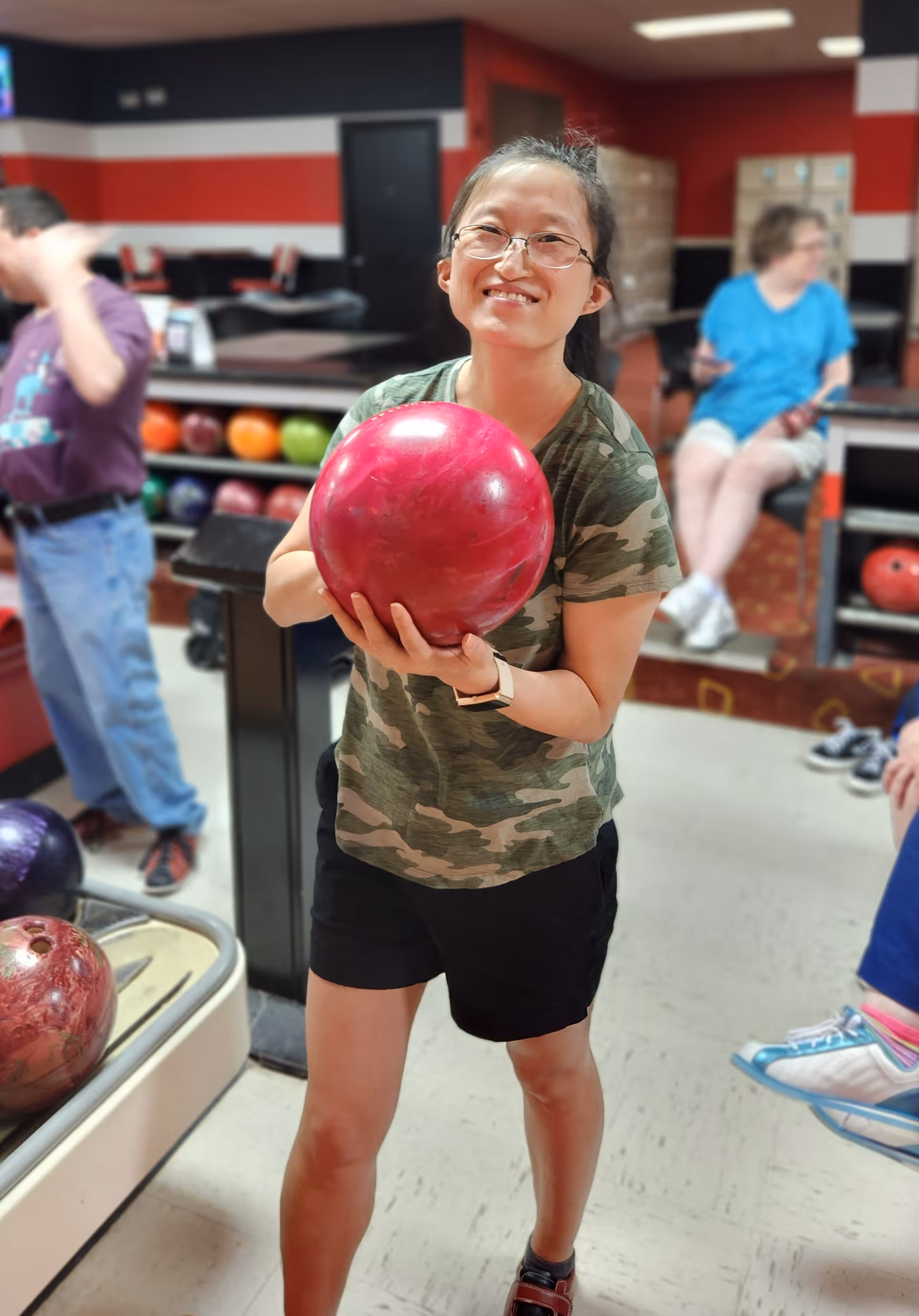 A smiling woman wearing glasses, a camouflage t-shirt, and black shorts is holding a pink bowling ball in a bowling alley. In the background, there are other people, bowling balls on racks, and seating areas with red and black walls.