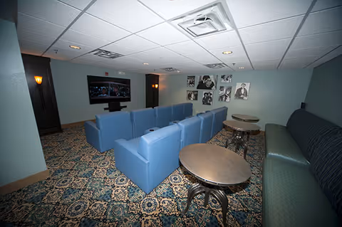 A cozy media room with a patterned carpet, blue cushioned chairs arranged in two rows facing a wall-mounted flat screen TV, and a long cushioned bench along the right wall. The walls are decorated with black and white photos, and there are small round tables in front of the bench. The ceiling has recessed lighting and air vents.