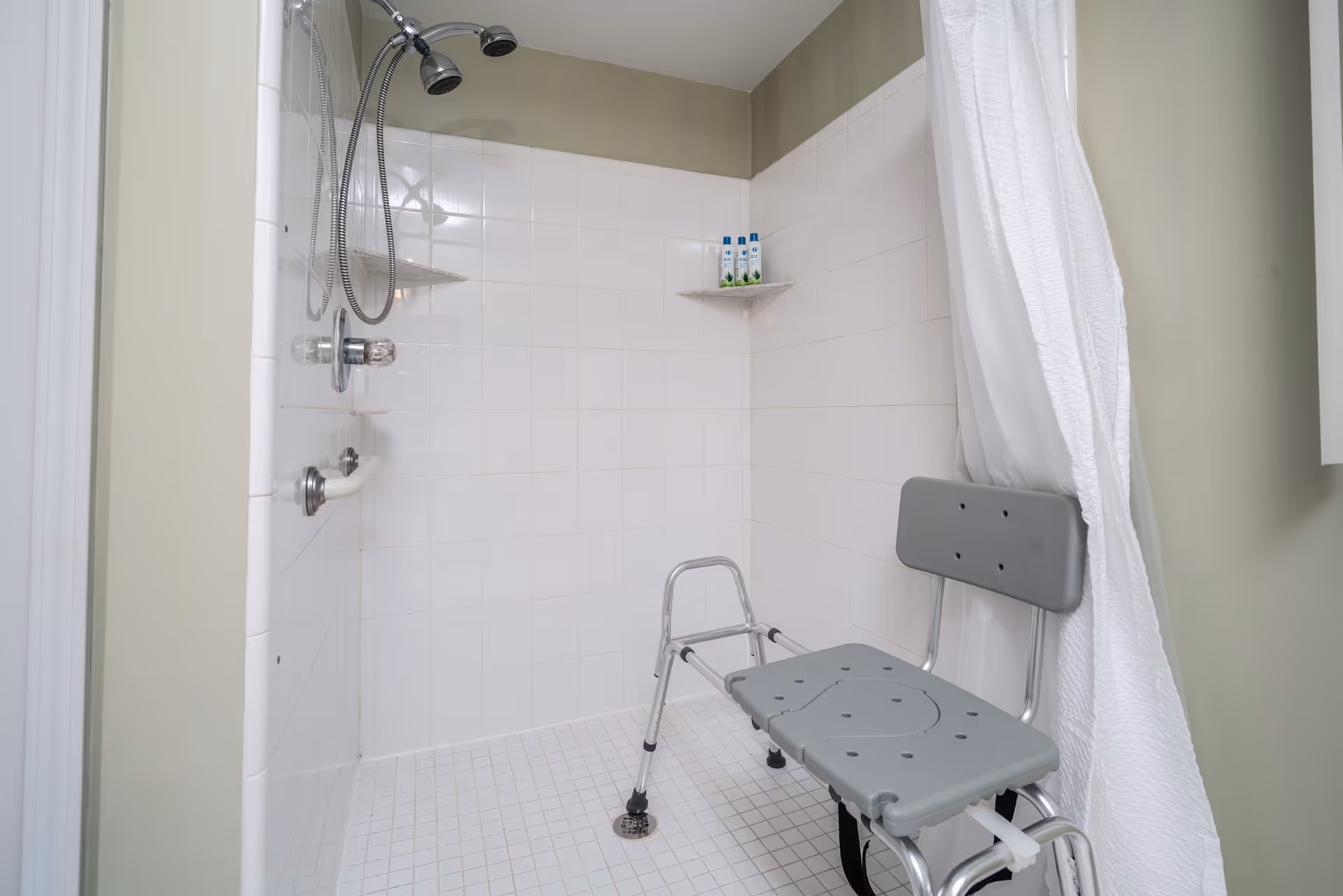 A tiled shower area with a white curtain partially open, featuring a gray shower chair with backrest, a handheld showerhead, grab bars, and three bottles of shampoo or body wash on a corner shelf.
