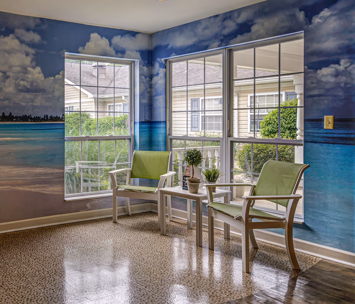 A small seating area with two green chairs and a white table between them, decorated with small potted plants. The walls are painted with a mural depicting a beach scene with blue sky, clouds, and ocean. Large windows let in natural light and show the exterior of the building and greenery outside.