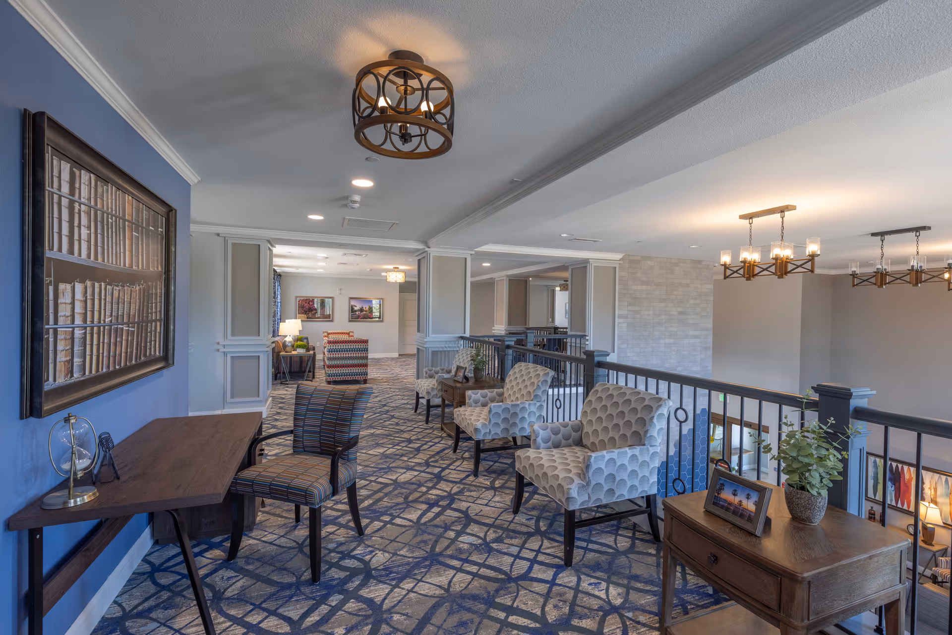 Interior view of a senior living facility hallway with patterned carpet, several upholstered armchairs, a wooden desk with a chair, framed artwork on the walls, and modern ceiling light fixtures. The space overlooks a lower level through a black metal railing.