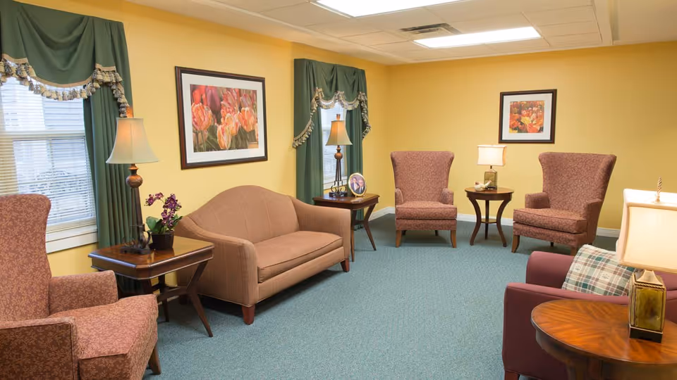 A cozy living room with yellow walls and green carpet featuring a brown sofa, four upholstered armchairs in a reddish pattern, three wooden side tables with lamps, green curtains with decorative trim on two windows, and framed floral artwork on the walls.