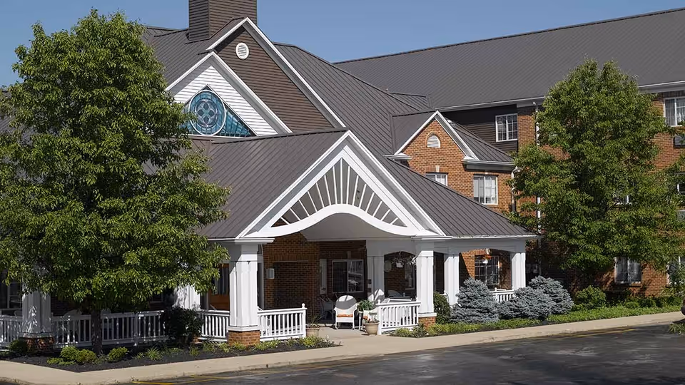 Exterior view of a senior living facility with a covered entrance featuring white columns and railings, surrounded by green trees and shrubs, with a brick building and multiple windows in the background under a clear sky.