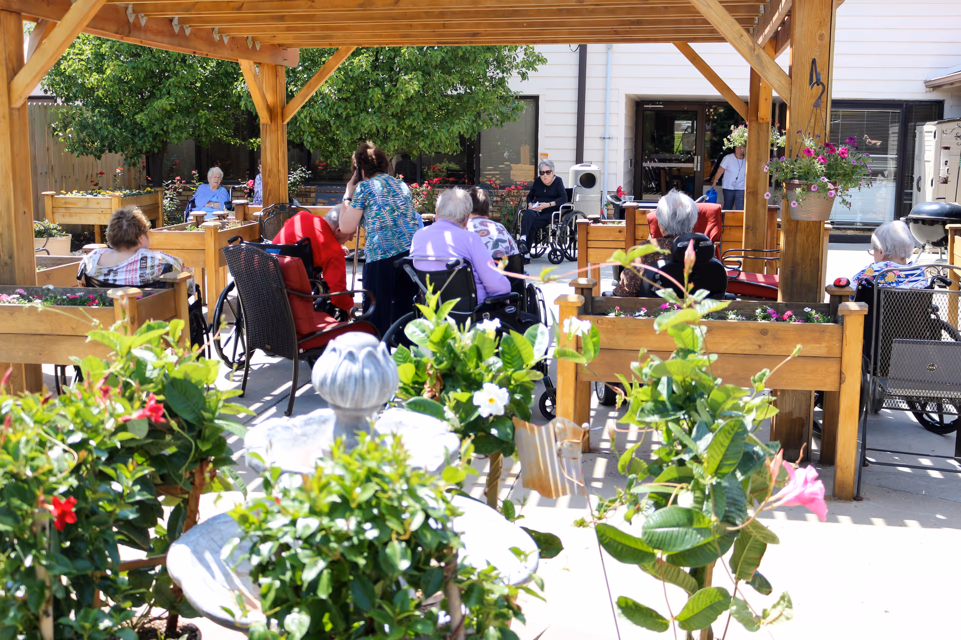 Elderly residents and staff seated and conversing under a wooden pergola in a flower-filled outdoor courtyard.