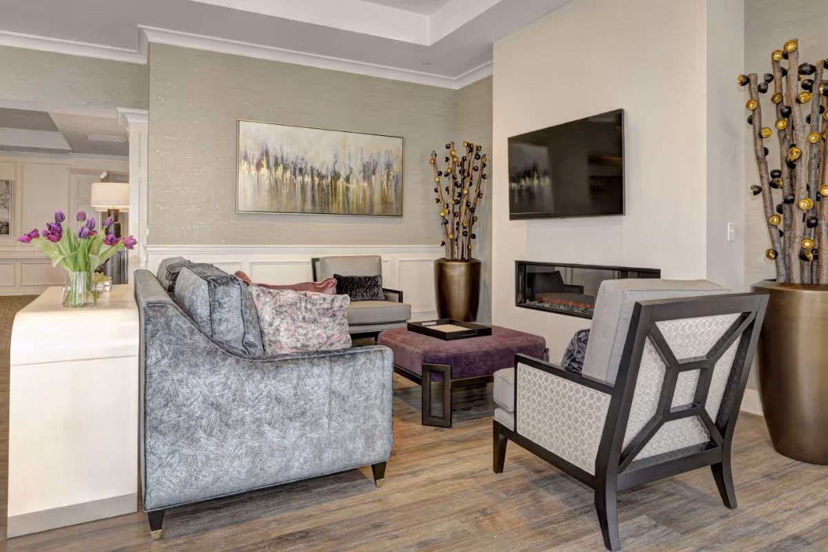 Stylish lounge with sofas and armchairs arranged around a central ottoman, a wall-mounted TV above a fireplace, artwork, and decorative plants.