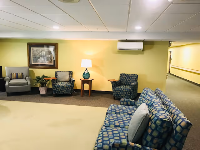A cozy seating area in a senior living facility with patterned blue armchairs and a matching sofa arranged around a small wooden table with a lamp. The walls are painted yellow, and there is a framed picture hanging on the wall. A hallway extends to the right.