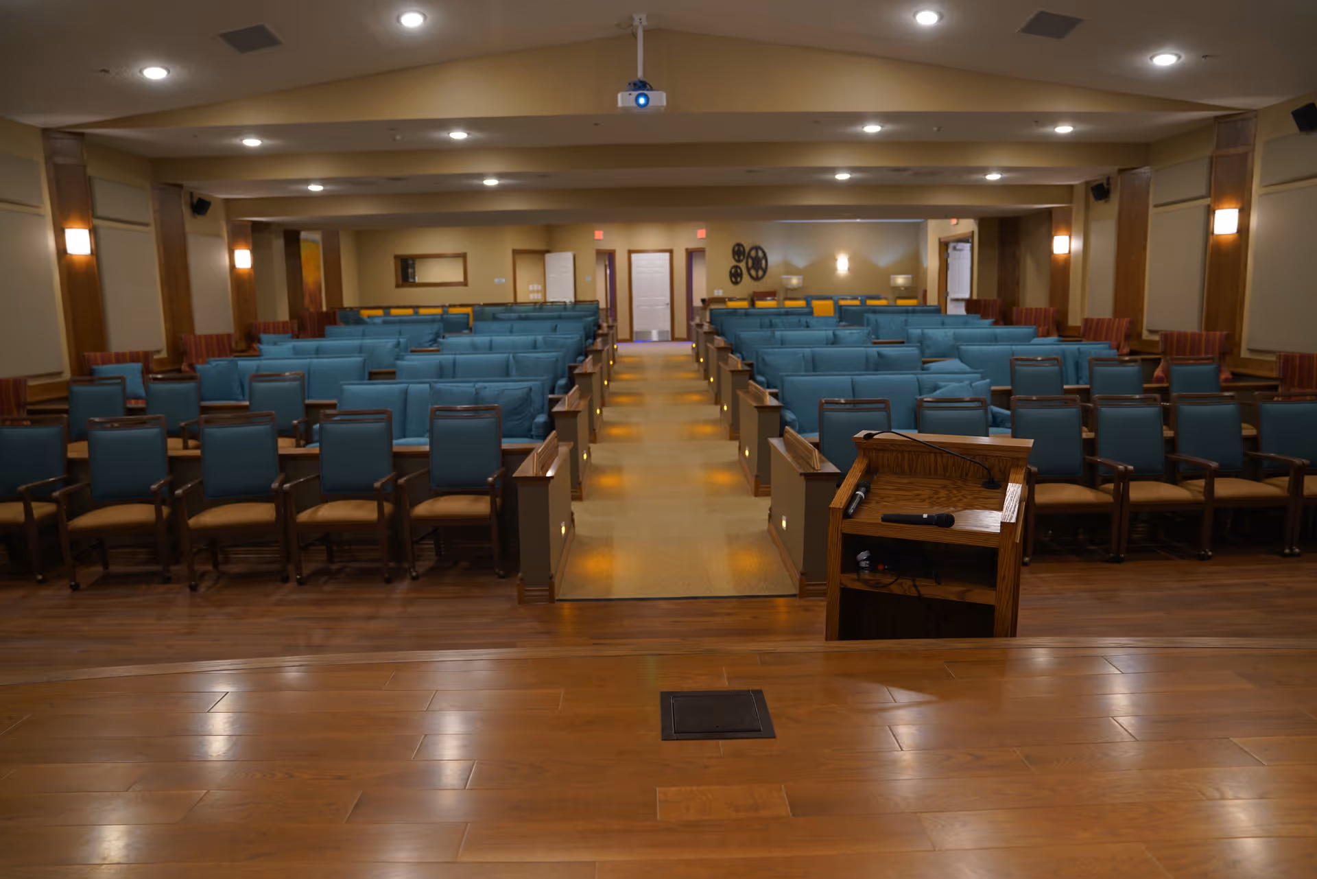 View from a wooden stage looking out over a large room with rows of blue cushioned chairs and armchairs arranged in an auditorium or meeting room style. There is a wooden podium with a microphone on the stage, and the room is softly lit with wall sconces and ceiling lights. The walls are beige with wooden panel accents.