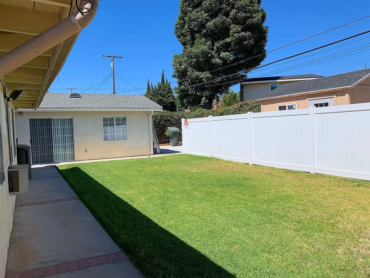 Sunny fenced backyard with a green lawn, concrete walkway, and a small outbuilding with a sliding glass door.