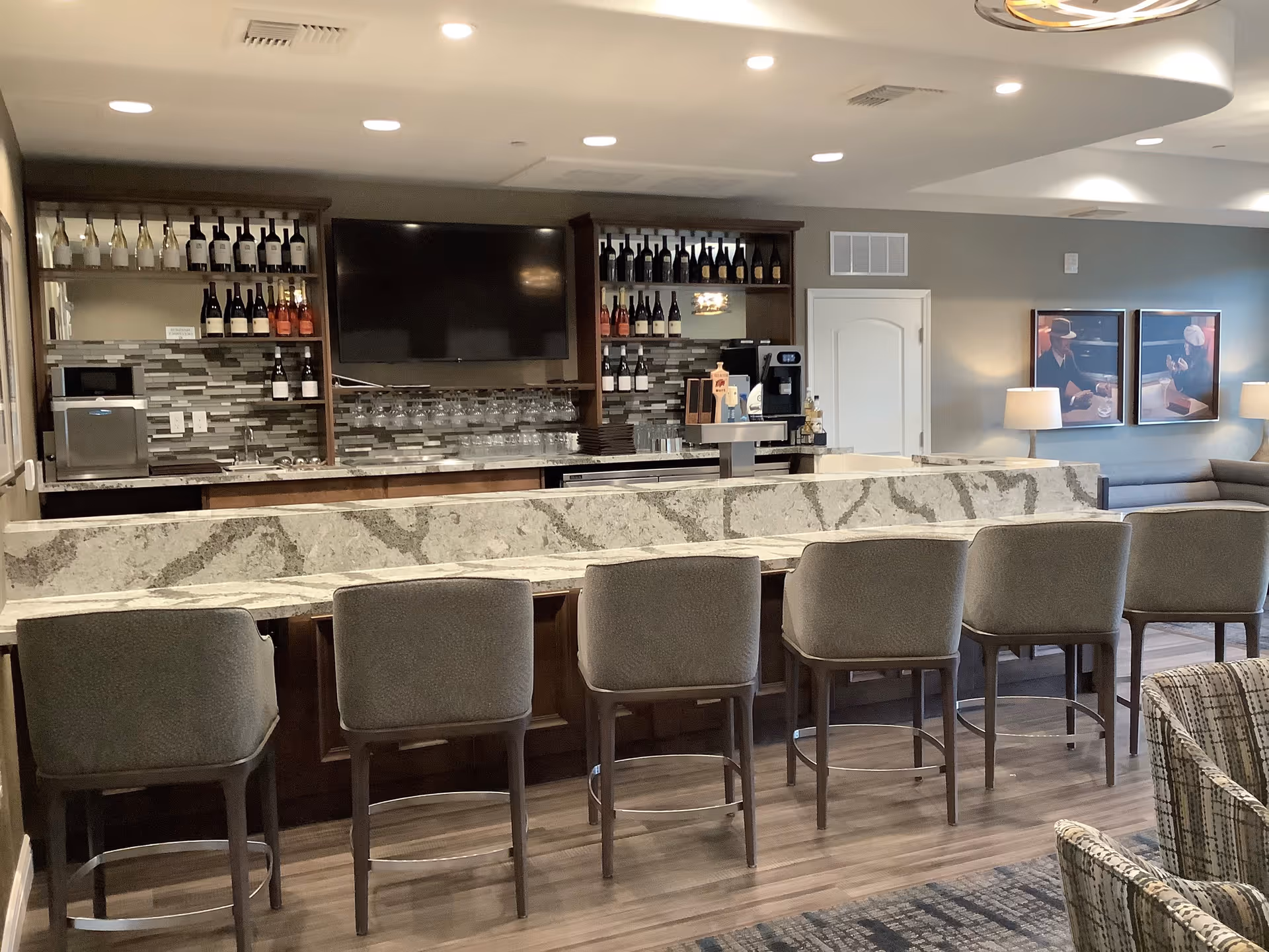 A modern bar area with a marble countertop and six upholstered bar stools. Behind the bar, there are shelves stocked with wine bottles and glasses, a large flat-screen TV mounted on the wall, and a coffee machine. The room has wood flooring, neutral-colored walls, and framed artwork with lamps on a side table in the background.