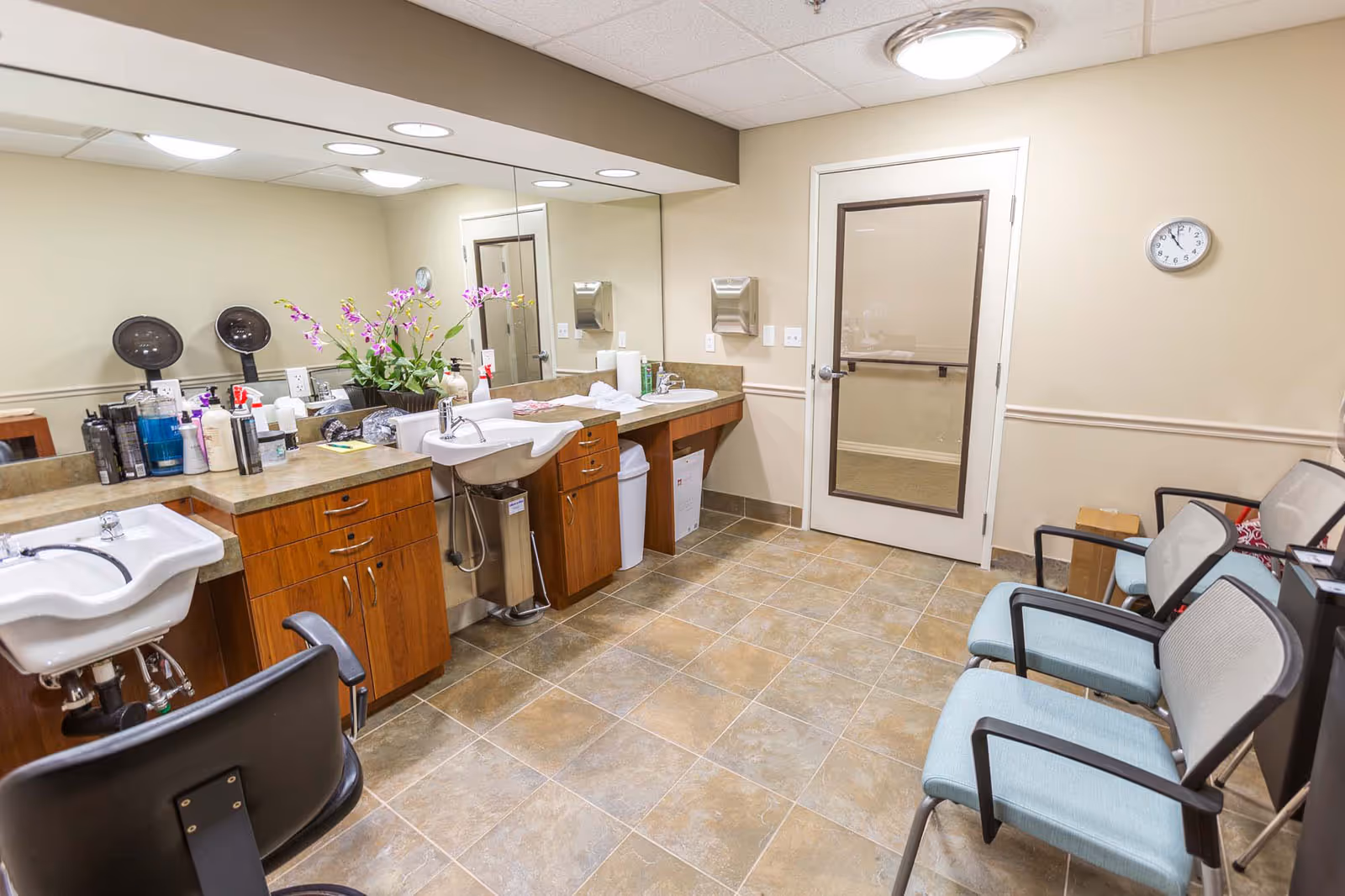 Interior of a salon or grooming room with two sinks, wooden cabinets, a large mirror, various hair care products on the counter, and a row of chairs along the right wall. The room has tiled flooring, beige walls, and a door with a window. A clock is mounted on the wall above the chairs.