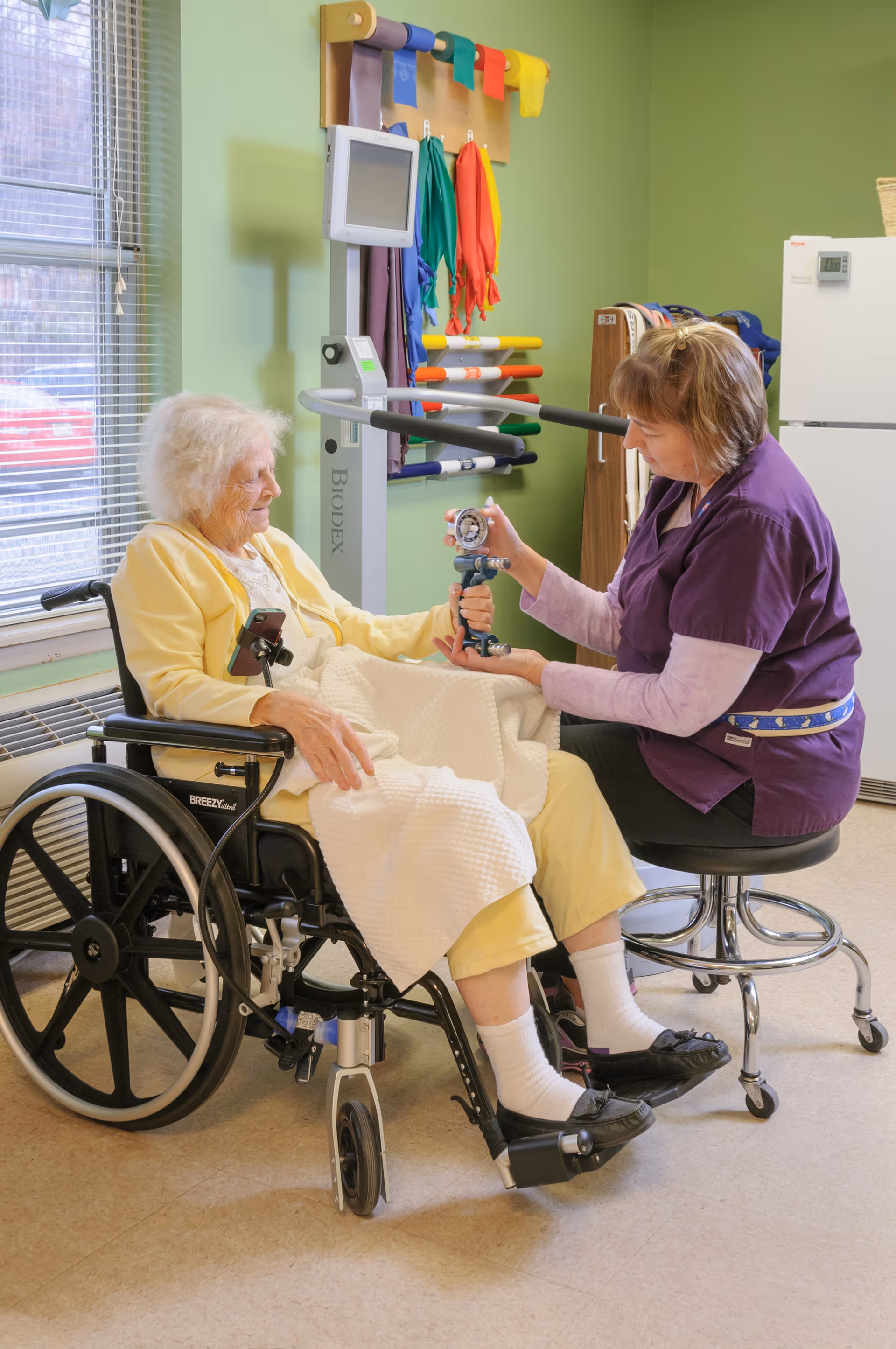 An elderly woman in a wheelchair is receiving physical therapy from a healthcare worker in a purple uniform. The therapist is holding a hand dynamometer to measure the woman's grip strength. The room has green walls, a window with blinds, and various colorful exercise bands hanging on the wall.