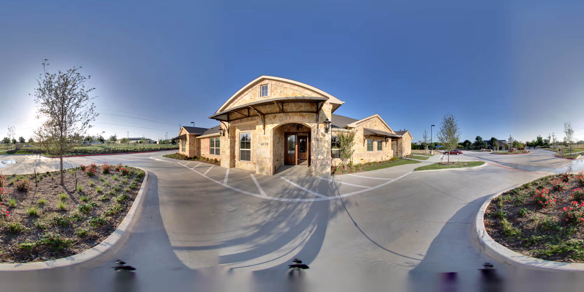 Exterior view of a single-story stone building with a covered entrance and surrounding landscaped areas including young trees and flower beds. The building is situated along a curved driveway with parking spaces and a clear blue sky overhead.