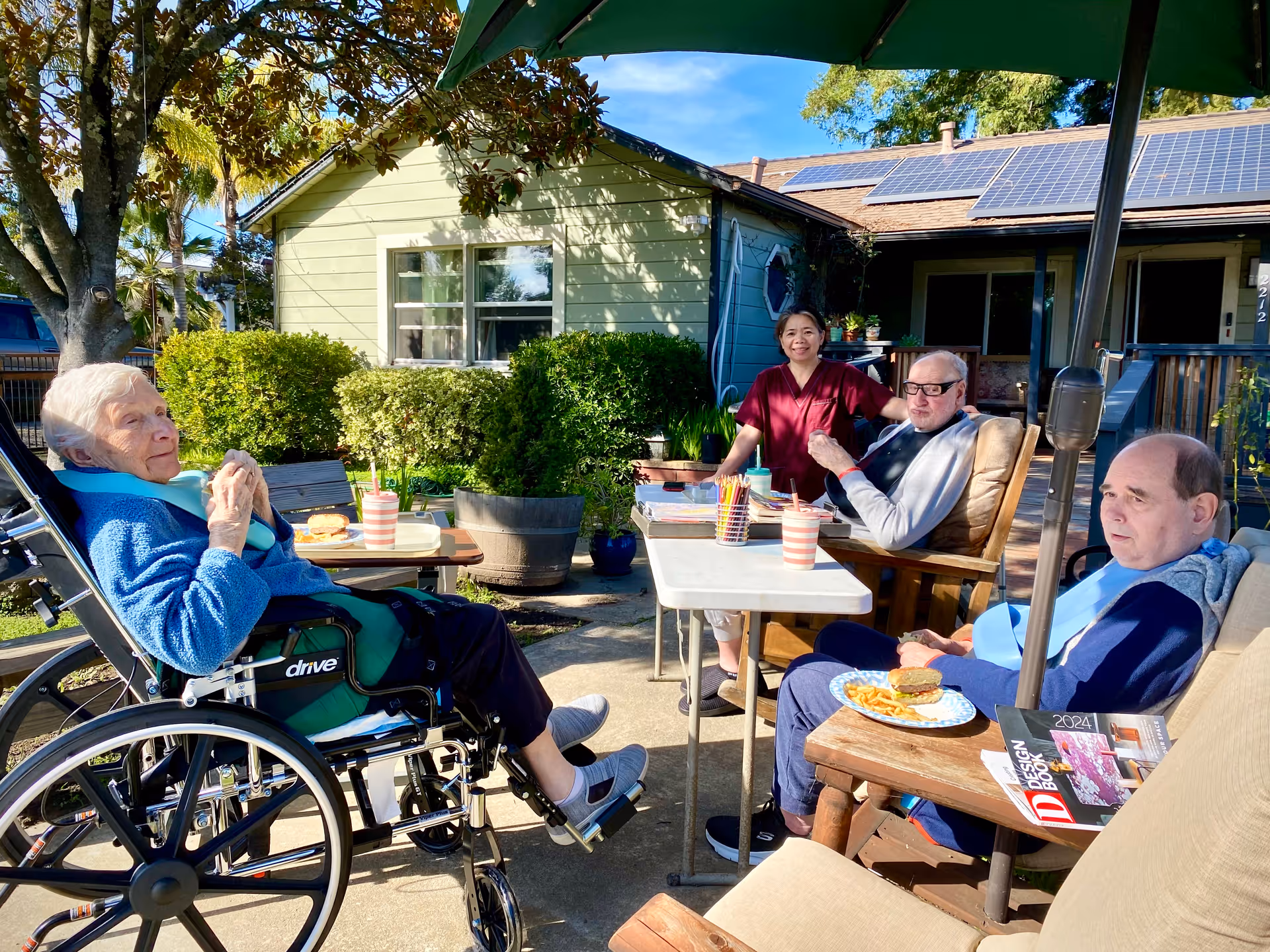 Three elderly individuals and a caregiver sitting outdoors in a garden area of a senior living facility. One elderly woman is in a wheelchair wearing a blue sweater, while two elderly men sit in chairs with plates of food in front of them. The caregiver stands behind a table with art supplies and drinks. The background shows a green building with solar panels on the roof and lush greenery.