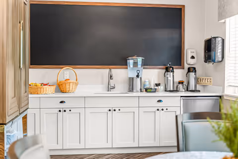 A clean and bright kitchenette area with white cabinets, a countertop holding a water dispenser, coffee thermoses, cups, and baskets. Above the counter is a large empty blackboard framed in wood. To the right, there is a window with blinds partially open, letting in natural light.