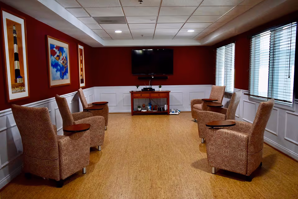 A cozy common room with six patterned armchairs arranged in two rows facing a wall-mounted flat screen TV. The room has red walls with white wainscoting, three framed pictures on the left wall, and three large windows with blinds on the right side. A wooden console table beneath the TV holds decorative items and electronic devices.