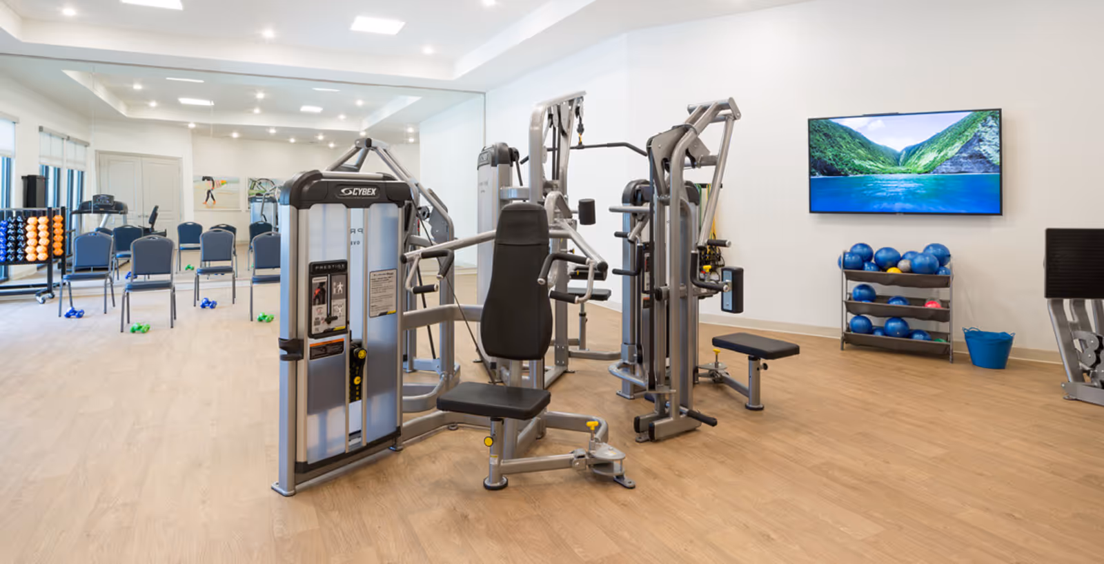 A bright fitness room with weight machines, free weights, exercise balls, chairs, and a wall-mounted TV.