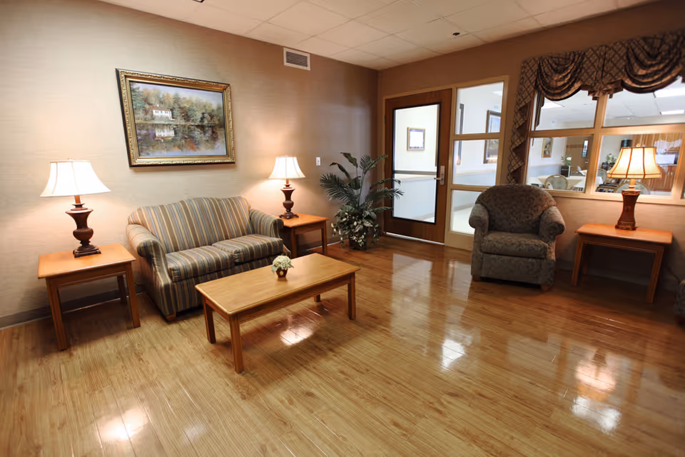 A cozy sitting area with a striped loveseat, a patterned armchair, two wooden side tables each with a lamp, a wooden coffee table with a small flower arrangement, a potted plant in the corner, a framed painting on the wall, and a door with glass panels next to a window with draped curtains.