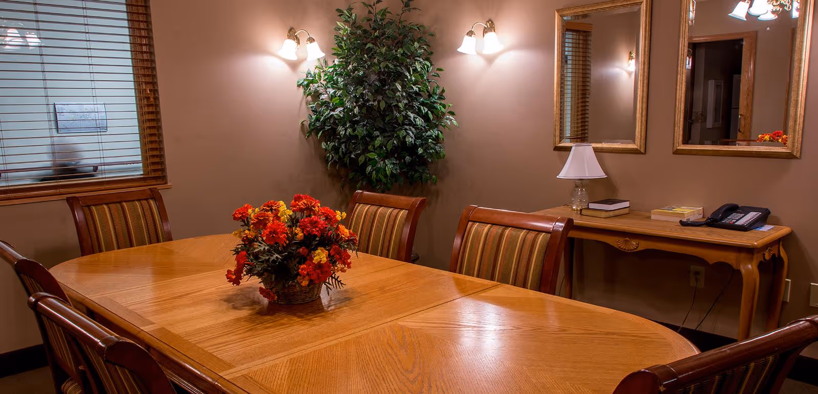 Wooden dining/meeting room with a long table and chairs, a floral centerpiece, wall sconces, mirrors, a potted plant, and a console table with a lamp and phone.