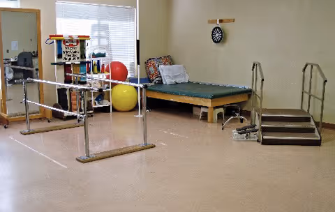 Physical therapy room with parallel bars for walking exercises, a padded therapy table with pillows, a small set of stairs with handrails, exercise balls, and shelves with various therapy equipment. A dartboard is mounted on the wall.