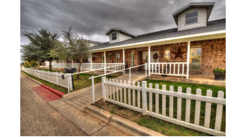 Exterior view of a single-story brick building with a white picket fence and a covered porch. The porch has railings and a ramp for accessibility. There are small trees and grass along the front, and the sky is overcast with gray clouds.