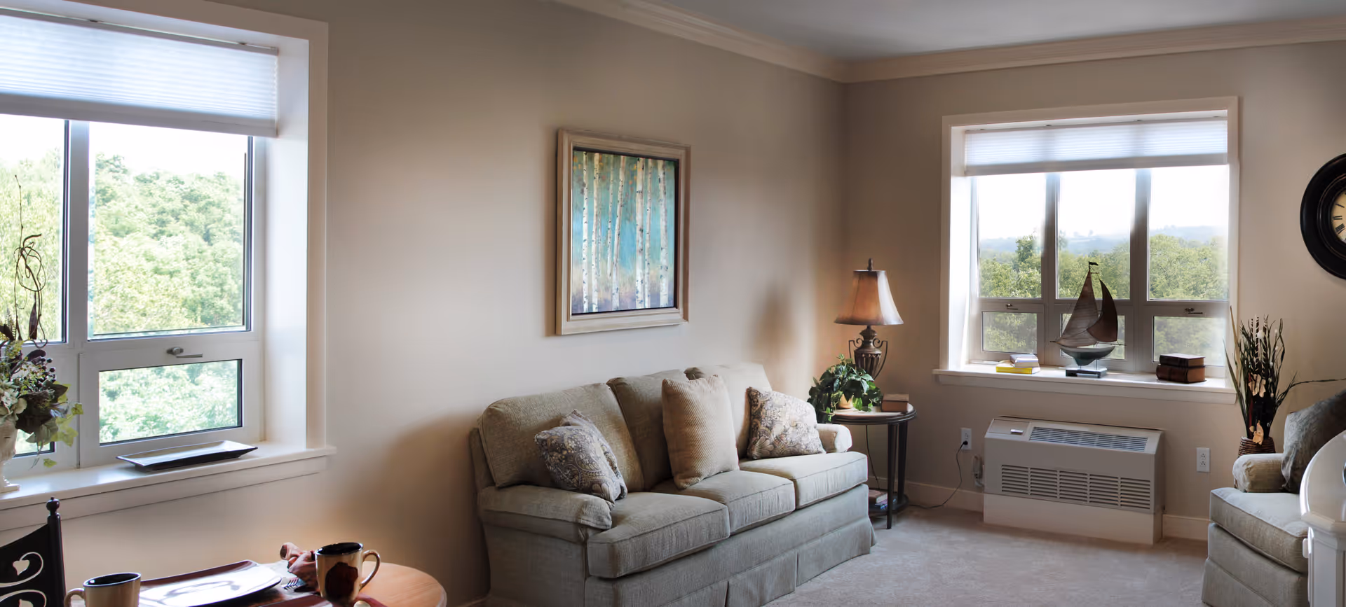 A cozy living room with a beige sofa adorned with patterned cushions, a side table with a lamp and plant, large windows with white blinds letting in natural light, a decorative sailboat on the windowsill, and a framed painting of birch trees on the wall.