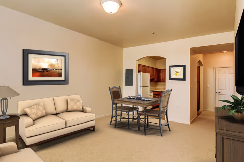 A cozy living area with a beige sofa and matching armchair, a wooden side table with a lamp, a small dining table with four chairs, and a kitchen with wooden cabinets and white appliances in the background. The walls are light-colored with framed artwork, and there is a ceiling light fixture.